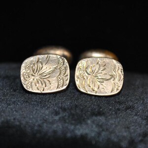 May include: A pair of gold-toned cufflinks with a floral design. Each cufflink is square with rounded corners and features an engraved botanical pattern. The background is dark, highlighting the cufflinks' intricate details and metallic sheen.