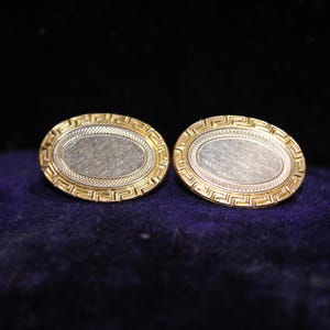 May include: A pair of gold-toned oval cufflinks with a silver-colored center. The gold border features a Greek key design. The cufflinks are displayed on a dark blue surface.