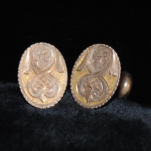 May include: A pair of antique gold-toned cufflinks. Each oval cufflink features an engraved design with a letter "A" and a heart-shaped element. The background has a gold color.
