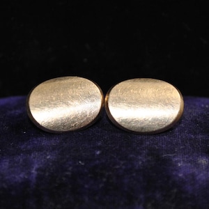 May include: A pair of oval-shaped cufflinks with a brushed gold-tone finish. The surface of each cufflink features a subtle, directional texture that catches the light, creating a soft metallic sheen across the rounded, smooth edges of the metal.