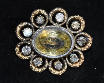 May include: A vintage brooch with a central oval yellow gemstone surrounded by smaller clear gemstones and gold-toned metalwork. The brooch has a floral design with a silver-toned border around the central stone. The background is black.