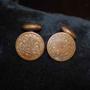 May include: A pair of round, metallic cufflinks with a warm, aged bronze finish. Each cufflink features a circular face with an intricate, engraved pattern consisting of central radiating lines surrounded by a decorative border of small, repeating shapes.