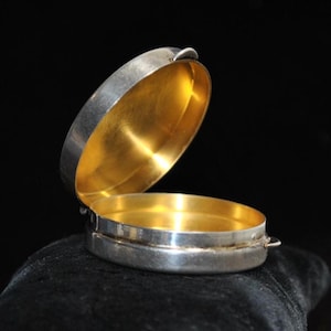 May include: An open, oval-shaped silver box with a gold interior. The lid is slightly ajar, revealing the gold lining. The box is set against a dark background, highlighting its metallic sheen. The box has a small loop on the side.