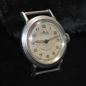 Pu&ograve; includere: Orologio da polso vintage Mido Multifort Super-Automatic con cassa color argento e quadrante color crema. L'orologio presenta numeri e lancette nere, e il logo Mido. Il quadrante &egrave; rotondo e ha una corona.