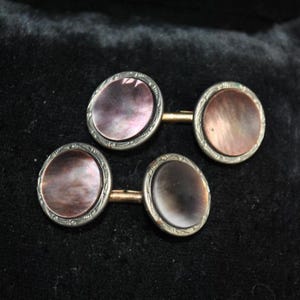 May include: A pair of vintage cufflinks with oval, iridescent mother-of-pearl centers. The cufflinks have a silver-toned, ornate border and gold-toned connectors. The background is a dark, textured fabric.