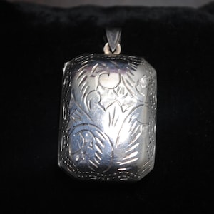May include: A rectangular silver pendant with an engraved floral design. The pendant has a bail at the top for a chain. The intricate pattern covers the front and sides of the pendant, creating a vintage aesthetic.