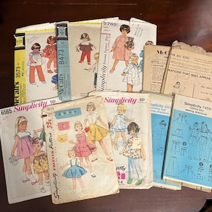 May include: A collection of vintage sewing patterns for children's clothing. The patterns feature illustrations of girls in various outfits, with text including "Simplicity" and "McCall's". The patterns are in various sizes and include instructions and cutting diagrams.