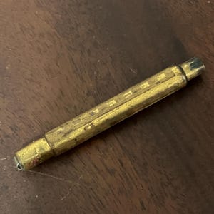 May include: A vintage brass pencil with a rectangular body and a slightly worn appearance. The pencil has a detailed, engraved design along its length and a small, dark tip. The pencil is lying on a dark wooden surface.