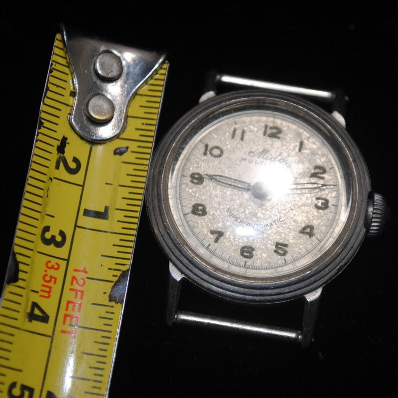 Pu&ograve; includere: Un orologio da polso vintage Mido Superautomatic con cassa color argento e quadrante chiaro e strutturato. L'orologio ha lancette e numeri neri. Un metro a nastro giallo &egrave; accanto all'orologio.