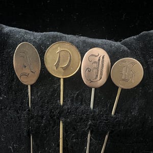 May include: Four antique monogram pins with ornate letter designs. The pins feature the letters A, D, J, and F, each in a unique script. The pins are made of a gold-toned metal and are set against a dark background.