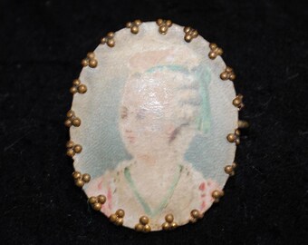 May include: Oval-shaped antique brooch featuring a portrait of a person with a hat and a green ribbon. The portrait is surrounded by a border of small, gold-colored beads. The background is a soft blue and pink.