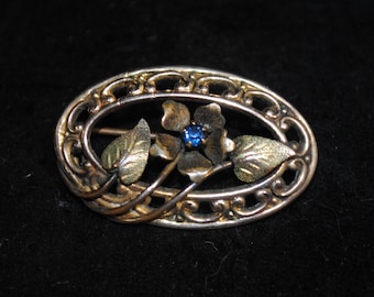May include: Oval-shaped vintage brooch with a floral design. The brooch features a central blue gemstone surrounded by gold-toned leaves and petals. The outer frame is silver-toned with an openwork design, creating an elegant and detailed accessory.