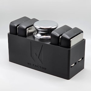 May include: A black rectangular holder featuring the text N1 FACTOR embossed on the front. The holder contains four black and silver rectangular blocks and one central silver circular knob on the top surface, all arranged in a structured row.