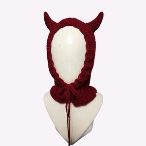 May include: A burgundy crocheted hood with two curved horns at the top. The hood has a neck warmer with a tie closure. The item is displayed on a white mannequin head against a white background.