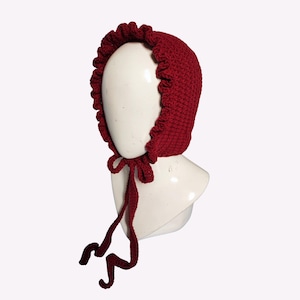 Puede incluir: Un gorro tejido de color burdeos con borde con volantes y cierre de lazo. El gorro se muestra en una cabeza de maniquí blanca. La textura tejida es visible y las cintas cuelgan. Adecuado para bebés y niños.