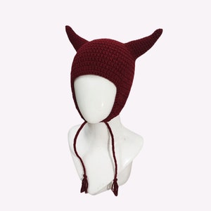 May include: A burgundy crocheted hat with two horns on top. The hat has a long tie that can be tied under the chin.