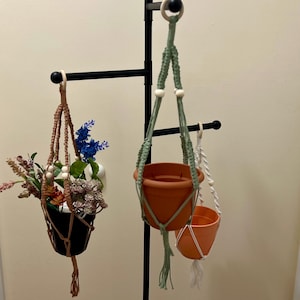 May include: Three macrame plant hangers with wooden beads hanging from a black metal stand. The hangers are in different colors: green, brown, and white. Each hanger has a terracotta pot.