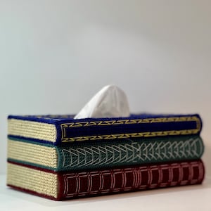 May include: A decorative tissue box shaped like a stack of books. The top book is navy blue with gold trim, the middle is teal with white stitching, and the bottom is burgundy. White tissues peek out from the top.
