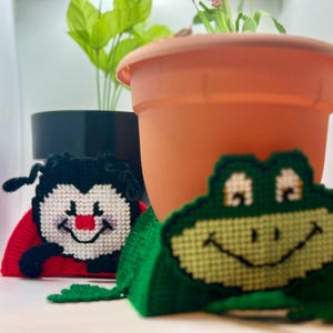 May include: Three handmade felt ornaments, a ladybug, a frog, and a butterfly, with embroidered details. The ornaments are sitting in front of a potted plant.