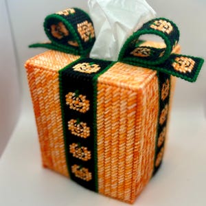 May include: Orange and green tissue box cover with a pumpkin design. The cover is made of plastic canvas and has a ribbon tied around it.