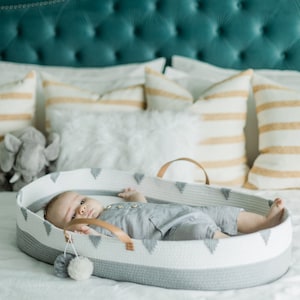 May include: A baby lies in a white and gray woven bassinet with a leather handle. The bassinet has a geometric design and is on a white bed. The baby is wearing a gray romper. Pillows and a stuffed elephant are in the background.