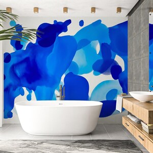 Op de afbeelding: Een moderne badkamer met een vrijstaand wit bad en een houten wastafel. De muur is voorzien van een levendig blauw abstract kunstontwerp. Een grote spiegel weerspiegelt de kunst en de wastafel. Een gevlochten mand en een groene plant zijn ook aanwezig.
