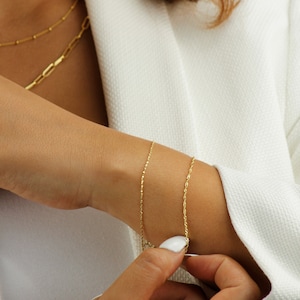 Op de afbeelding: Twee gouden kettingarmbanden om de pols van een persoon. De armband bovenop is een delicate ketting, en de armband onderaan is een dikkere touwketting.
