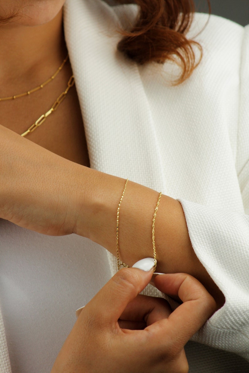 Op de afbeelding: Twee gouden kettingarmbanden om de pols van een persoon. De armband bovenop is een delicate ketting, en de armband onderaan is een dikkere touwketting.