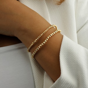 May include: Two gold beaded bracelets stacked on a person's wrist. The bracelets are made of small, round beads.