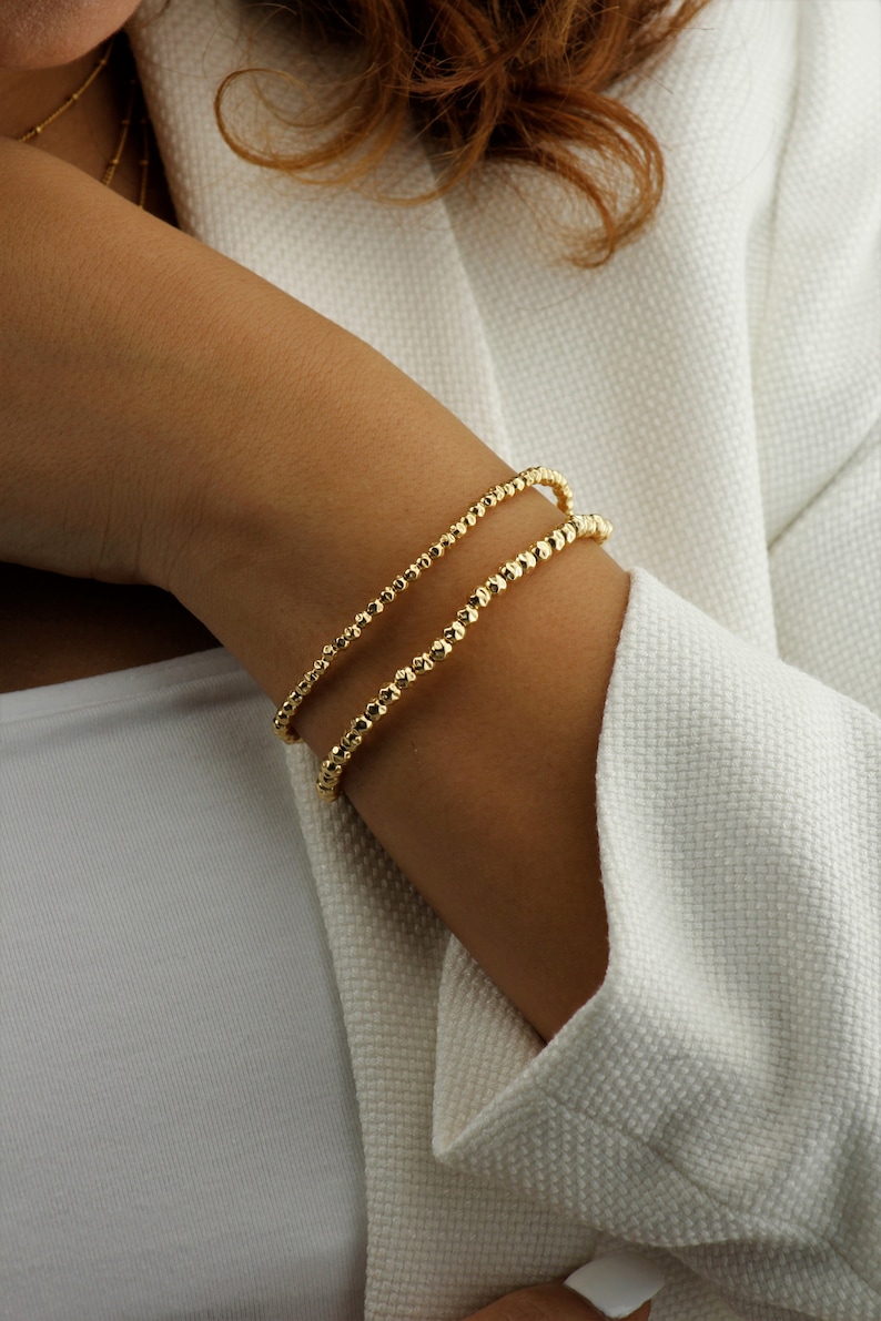 May include: Two gold beaded bracelets stacked on a person's wrist. The bracelets are made of small, round beads.
