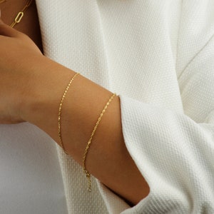 Op de afbeelding: Twee gouden kettingarmbanden om de pols van een persoon. De armbanden zijn allebei dun en delicaat, een is een touwketting en de andere een eenvoudigere ketting.