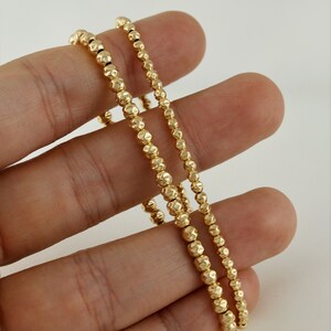 May include: Two gold beaded bracelets with a textured finish. The bracelets are stacked on top of each other and are being held by a hand.