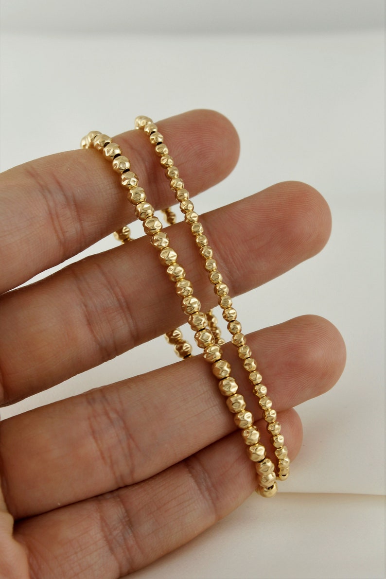 May include: Two gold beaded bracelets with a textured finish. The bracelets are stacked on top of each other and are being held by a hand.