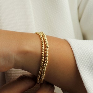 May include: Three gold beaded bracelets stacked on a person's wrist. The beads are small and round, giving the bracelets a delicate look.