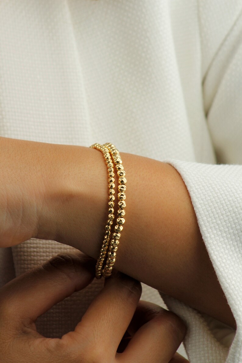 May include: Three gold beaded bracelets stacked on a person's wrist. The beads are small and round, giving the bracelets a delicate look.