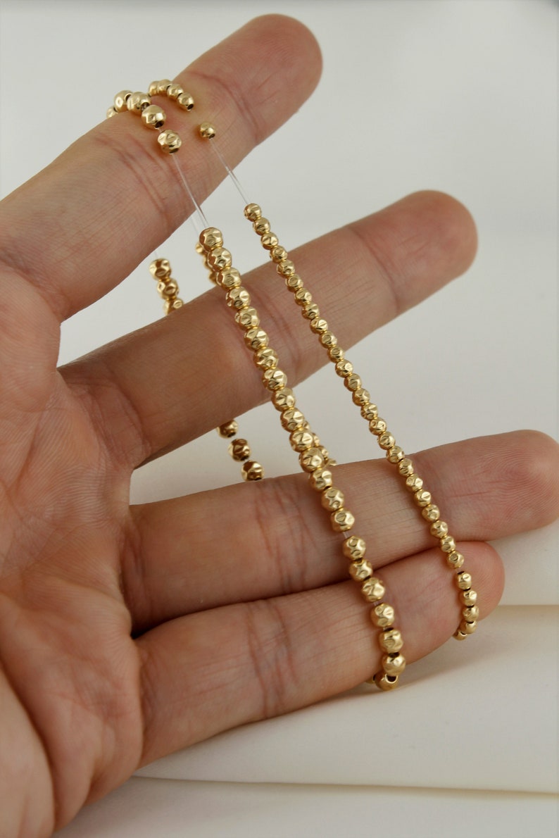 May include: Two gold beaded necklaces with a textured finish. The necklaces are on a hand and are both about 18 inches long.
