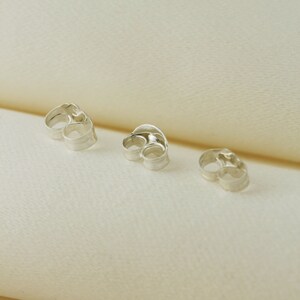 May include: Three silver earring backs, also known as earring stoppers, are shown on a white surface. The earring backs are designed to secure earrings to the earlobe.