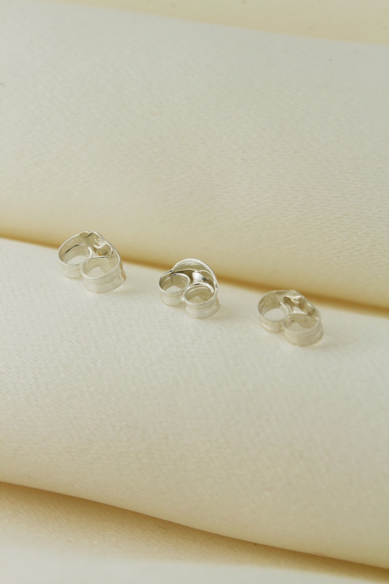 May include: Three silver earring backs, also known as earring stoppers, are shown on a white surface. The earring backs are designed to secure earrings to the earlobe.