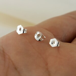 May include: Three silver earring backs, also known as earring stoppers, are shown on a hand. The backs are designed to secure earrings in place.