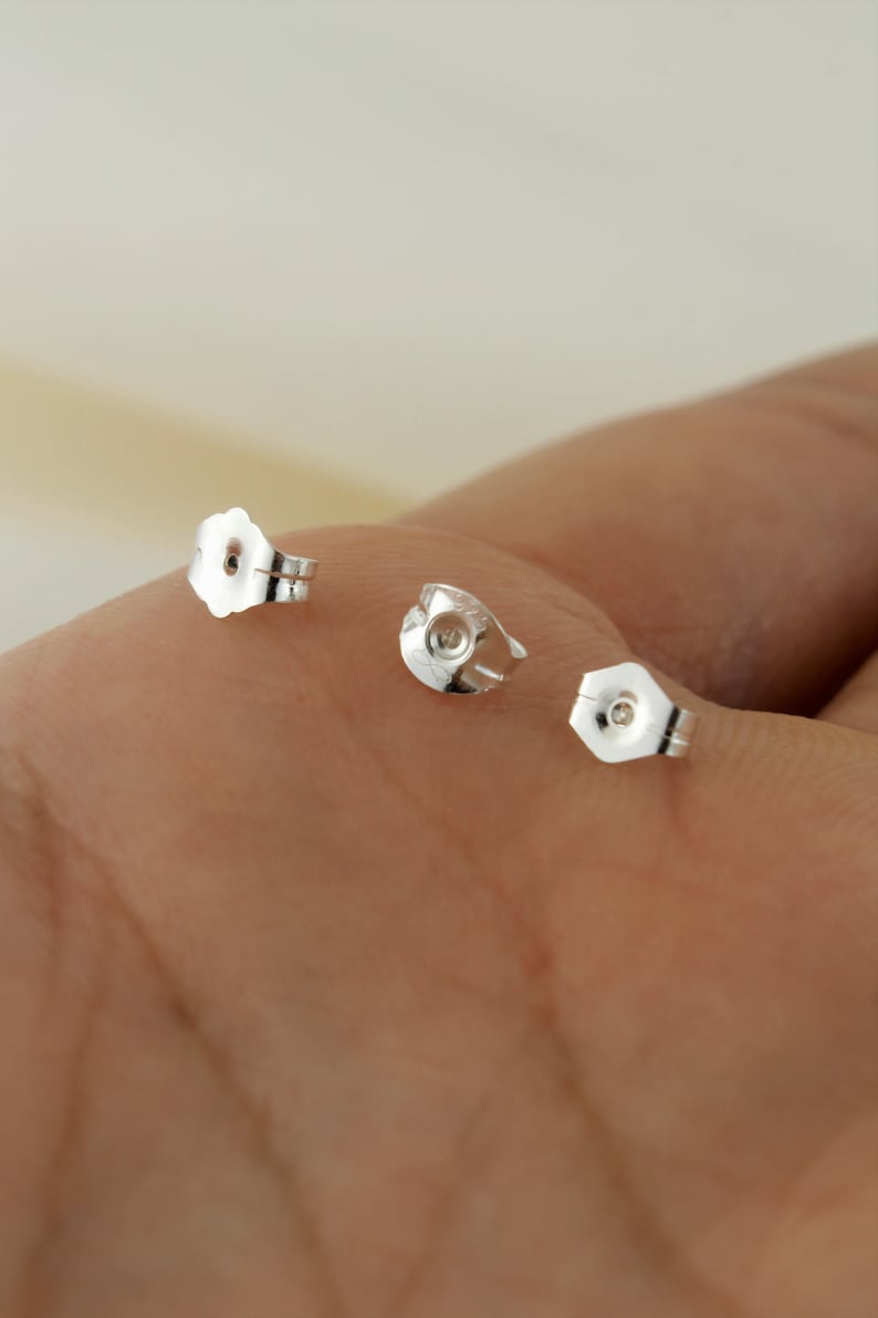 May include: Three silver earring backs, also known as earring stoppers, are shown on a hand. The backs are designed to secure earrings in place.