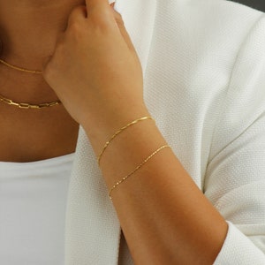 Op de afbeelding: Twee gouden kettingarmbanden om de pols van een persoon. De armbanden zijn dun en delicaat en zijn gemaakt van een eenvoudig kettingontwerp.