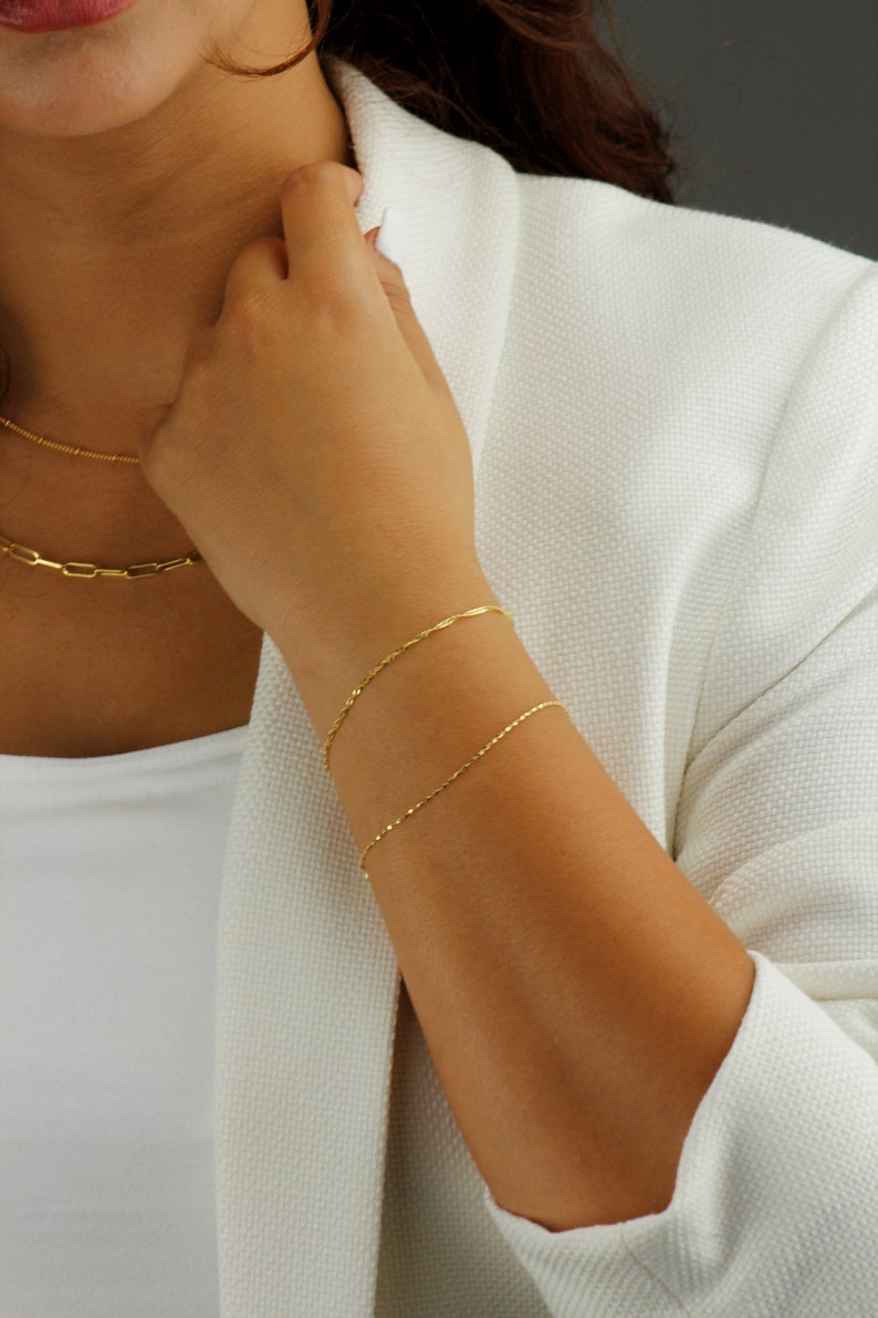 Op de afbeelding: Twee gouden kettingarmbanden om de pols van een persoon. De armbanden zijn dun en delicaat en zijn gemaakt van een eenvoudig kettingontwerp.