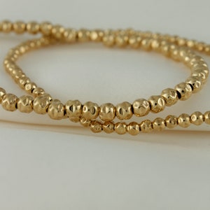 May include: Two gold beaded bracelets with a hammered texture. The bracelets are stacked on top of each other.