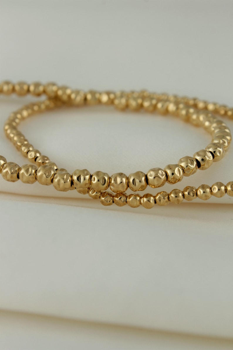 May include: Two gold beaded bracelets with a hammered texture. The bracelets are stacked on top of each other.