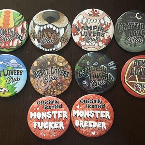 Pode incluir: Uma coleção de botões redondos com vários designs temáticos de monstros. Os botões apresentam textos como "Dragon Lovers Club", "Werewolf Lovers Club" e "Monster Fucker". Os botões são coloridos e têm um acabamento brilhante.
