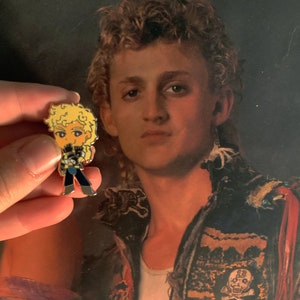 May include: A small, colorful enamel pin featuring a cartoon character with blonde curly hair, wearing a black and blue outfit. The pin is held in front of a portrait of a person with curly hair and a white shirt.