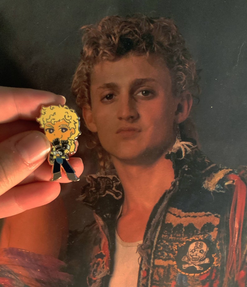 May include: A small, colorful enamel pin featuring a cartoon character with blonde curly hair, wearing a black and blue outfit. The pin is held in front of a portrait of a person with curly hair and a white shirt.