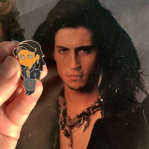 May include: A small enamel pin featuring a cartoon character with long black hair, a black jacket, and gray pants. The pin has orange skin and eyes with star-shaped highlights. The background is a vintage photograph of a man with long black hair.