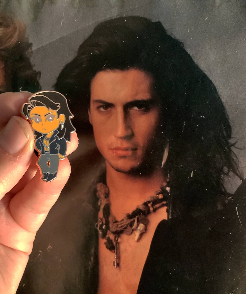 May include: A small enamel pin featuring a cartoon character with long black hair, a black jacket, and gray pants. The pin has orange skin and eyes with star-shaped highlights. The background is a vintage photograph of a man with long black hair.