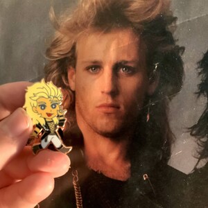 May include: A cartoon pin featuring a blonde character in a black and white outfit with gold accents. The pin is held in front of a vintage photograph of a man with blonde hair.